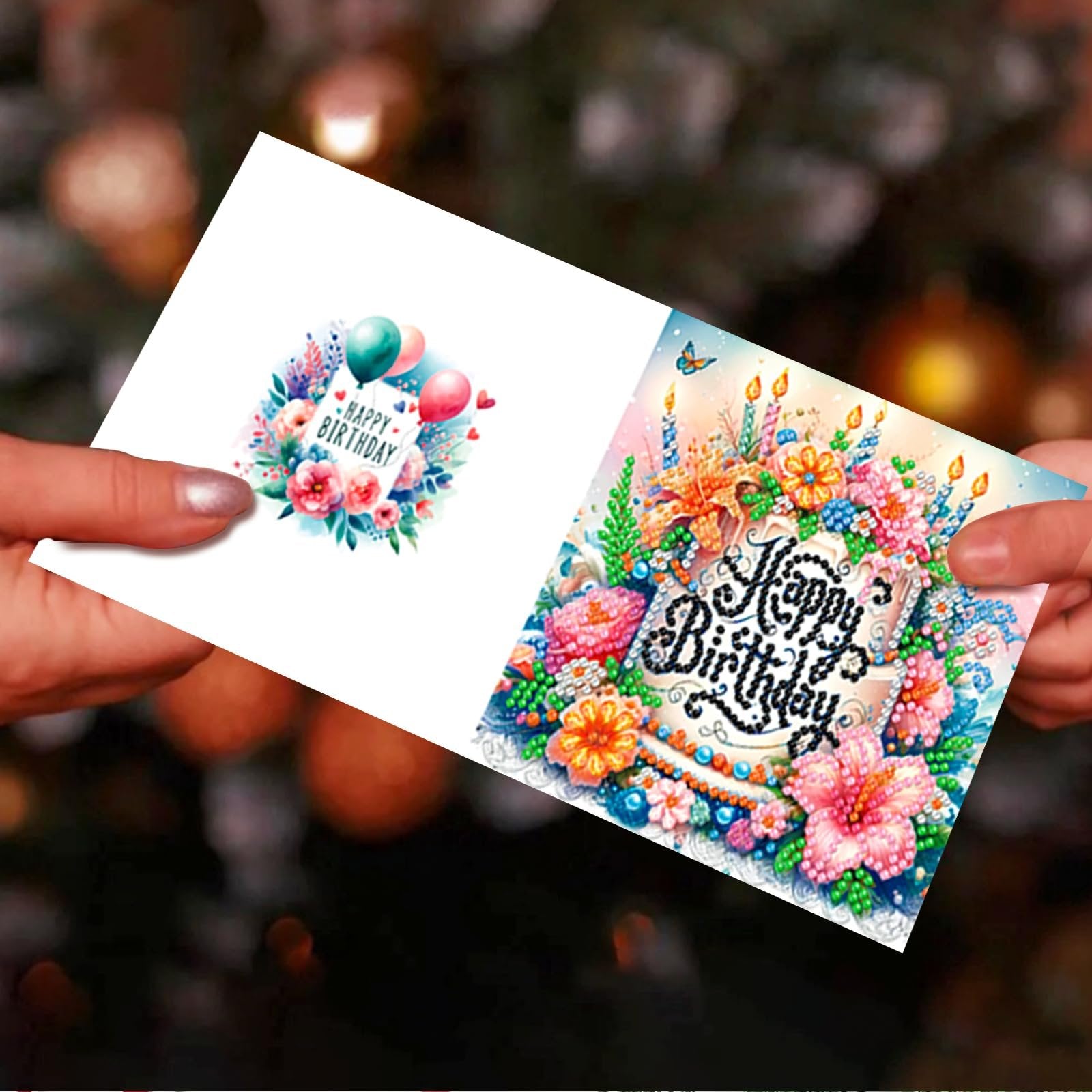 Two hands hold up diamond painting greeting cards with floral and balloon designs for birthdays.