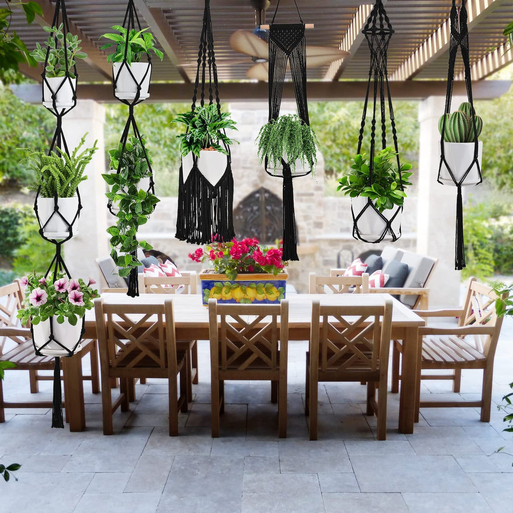 Six black macrame plant hangers with white pots and various plants hanging from a pergola over an outdoor dining set.