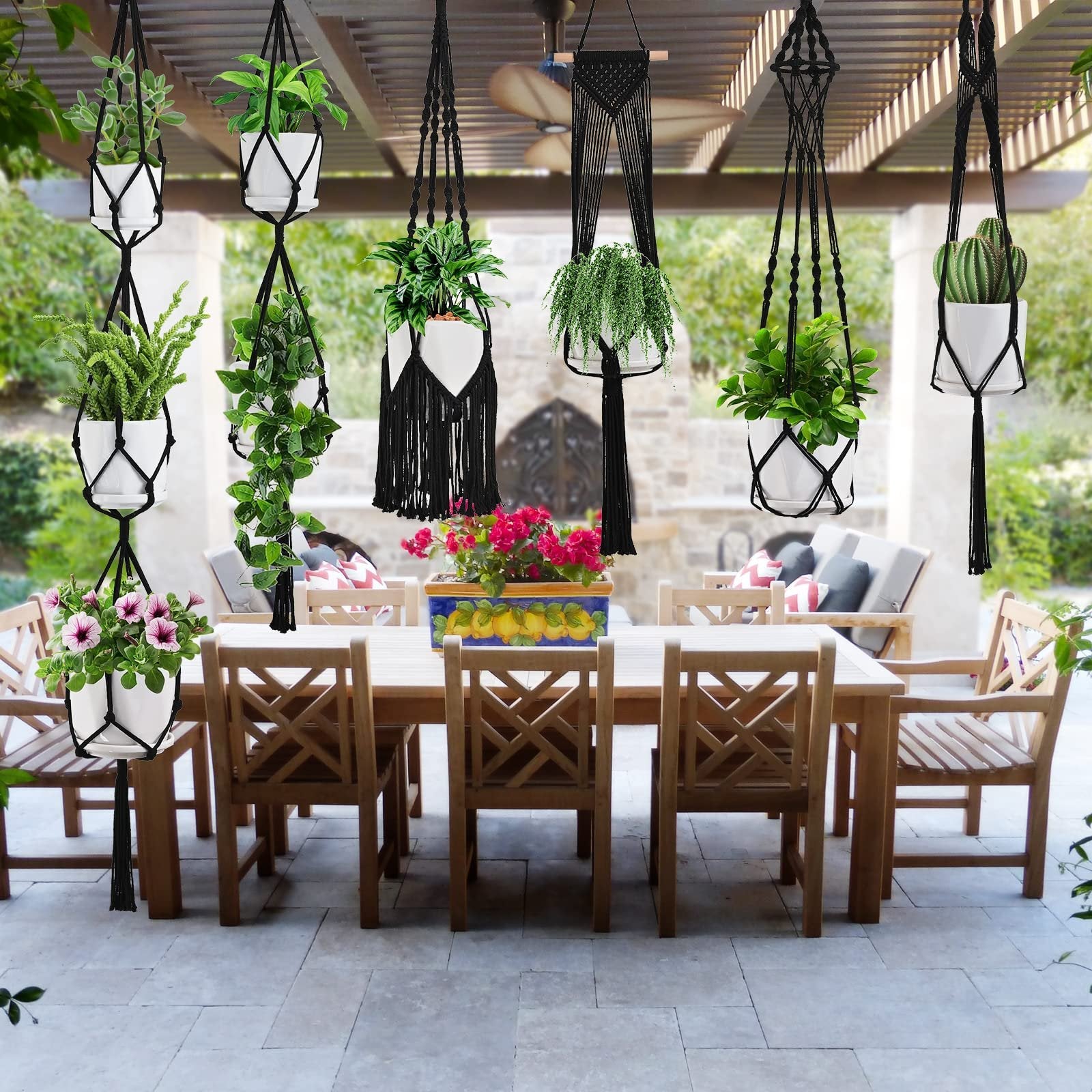 Six black macrame plant hangers with white pots and various plants hanging from a pergola over an outdoor dining set.