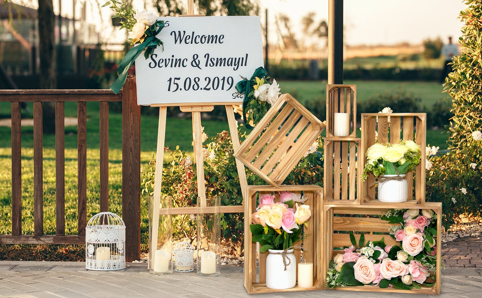 Wooden crates with floral arrangements and candles at an outdoor event with a welcome sign