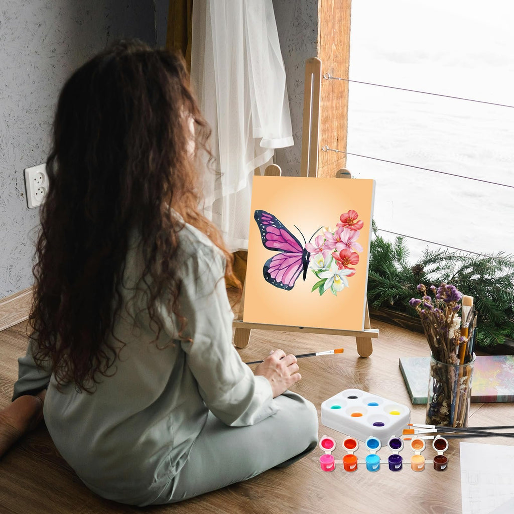 Woman painting a butterfly on canvas with acrylic paints and brushes laid out on the floor