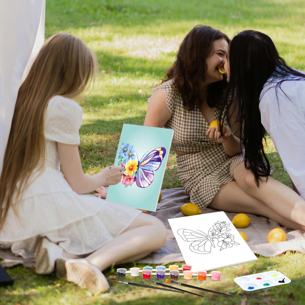 Three young women painting butterflies on canvas outdoors with acrylic paints and brushes laid out on a blanket.