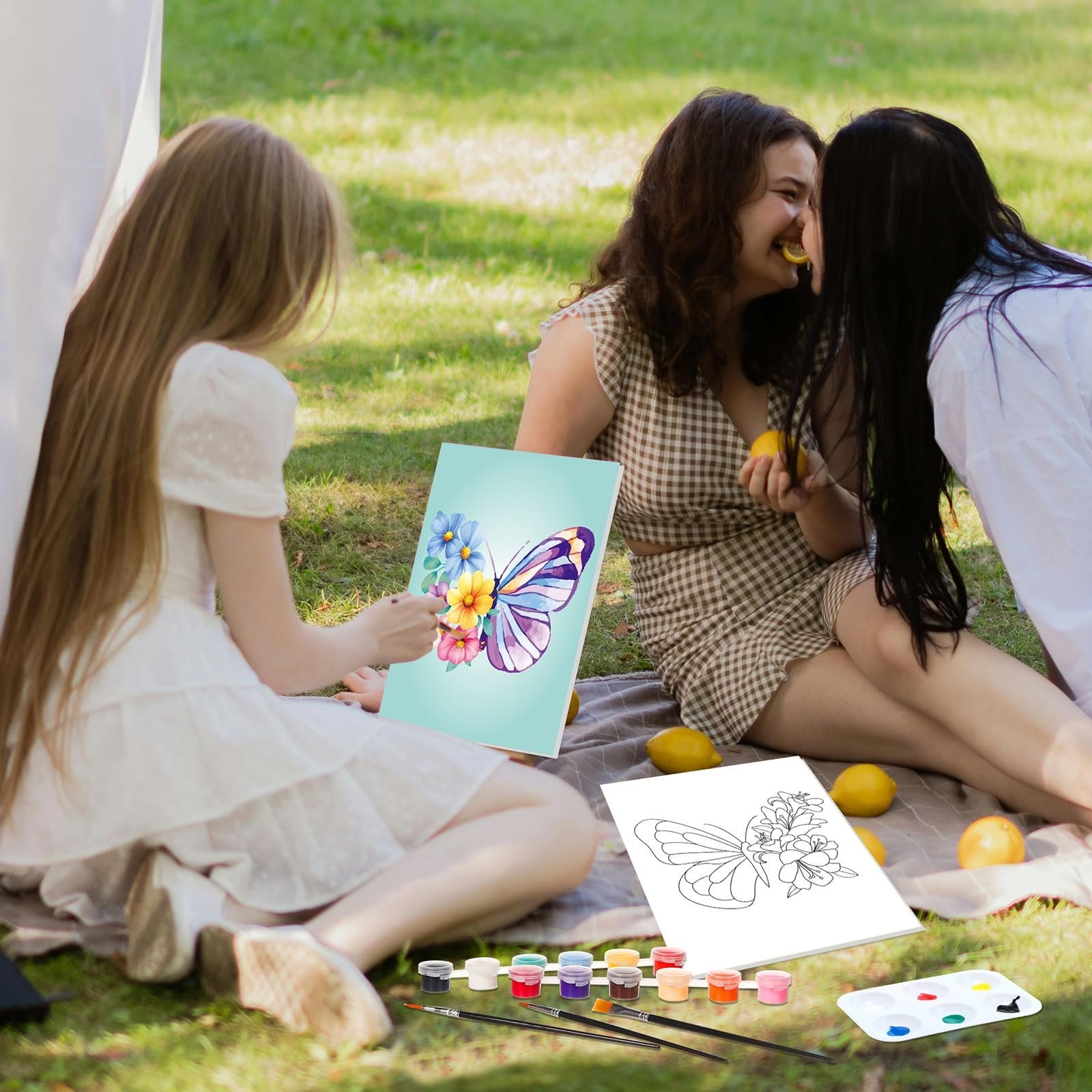 Three young women painting butterflies on canvas outdoors with acrylic paints and brushes laid out on a blanket.