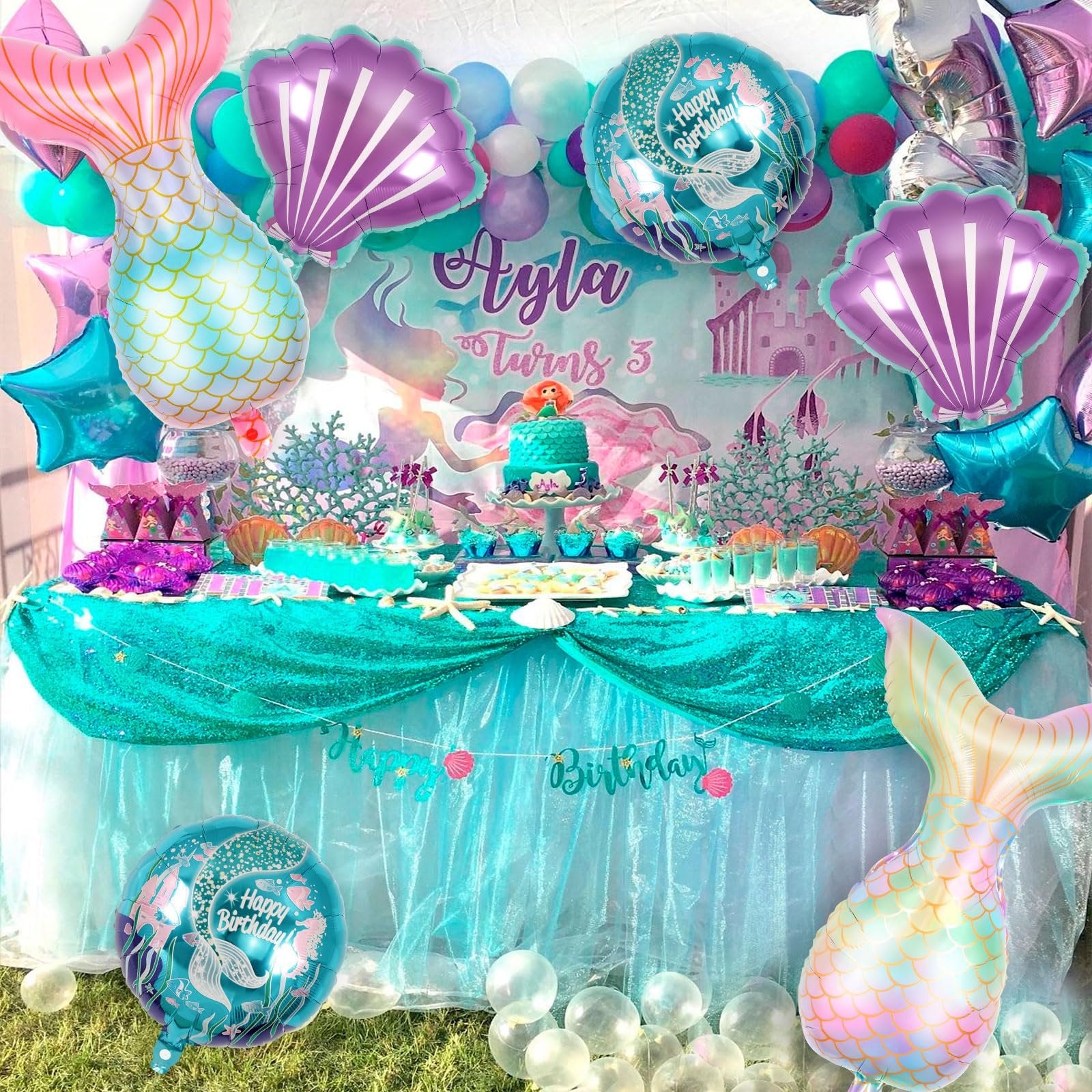 Mermaid themed birthday party table setting with balloons, cake, and cupcakes.