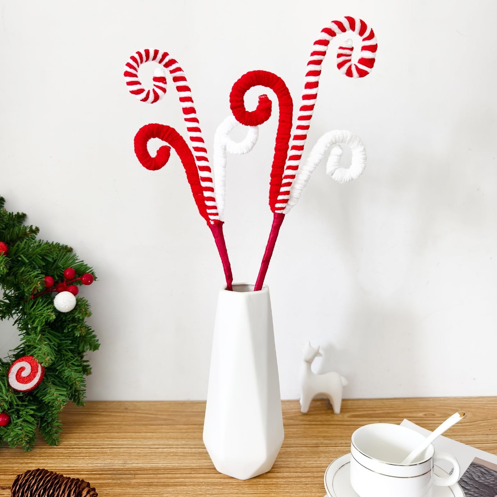 Six red and white candy cane picks in a white vase, with a Christmas wreath and pinecone on a wooden surface.
