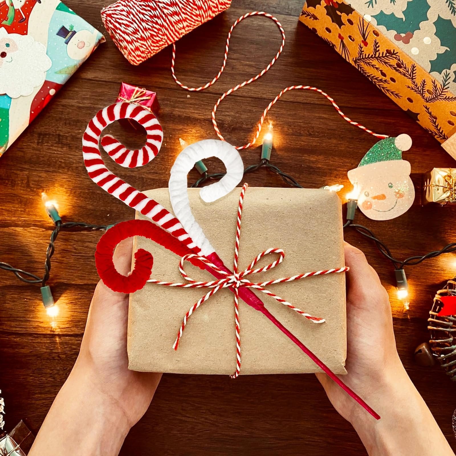 Hands holding a gift wrapped in brown paper, decorated with red and white candy cane picks and twine, surrounded by Christmas lights and wrapping paper.