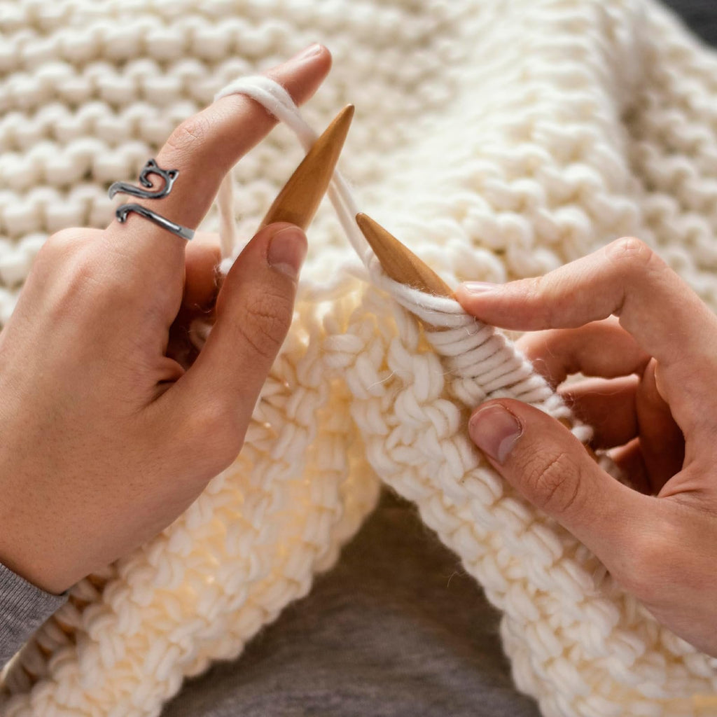 Close-up of hands knitting with white yarn and wooden needles, wearing a cat-shaped metal crochet ring on one finger.