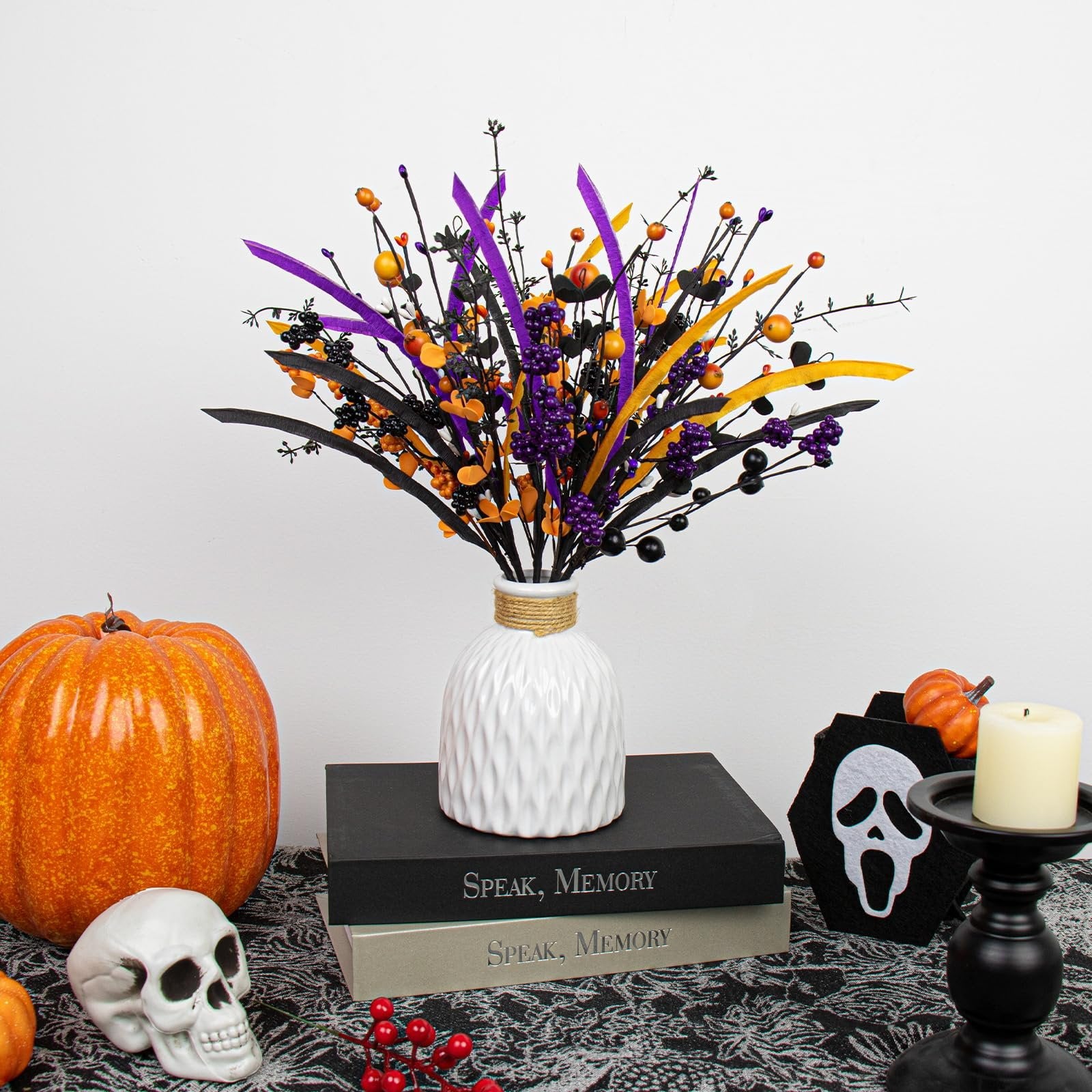 Halloween floral arrangement with black stems, orange berries, purple and yellow ribbons in a white vase, next to a pumpkin and skull.