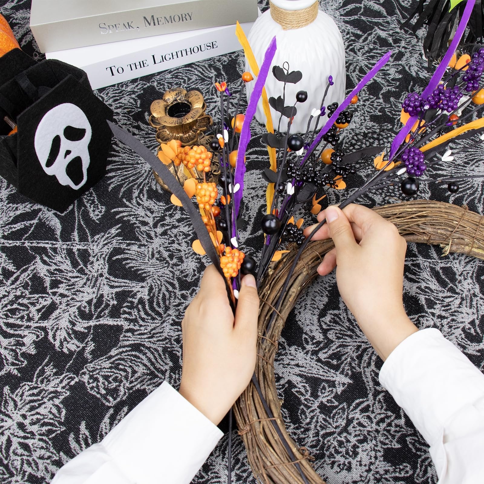 Hands arranging black floral picks with berries and orange accents onto a grapevine wreath