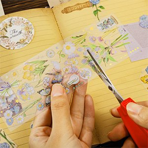Hands cutting a roll of decorative stickers featuring flowers and butterflies with scissors, next to an open notebook.