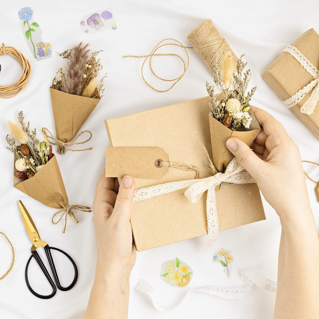 Hands wrapping a gift box with lace ribbon and a mini dried flower bouquet, surrounded by craft supplies.