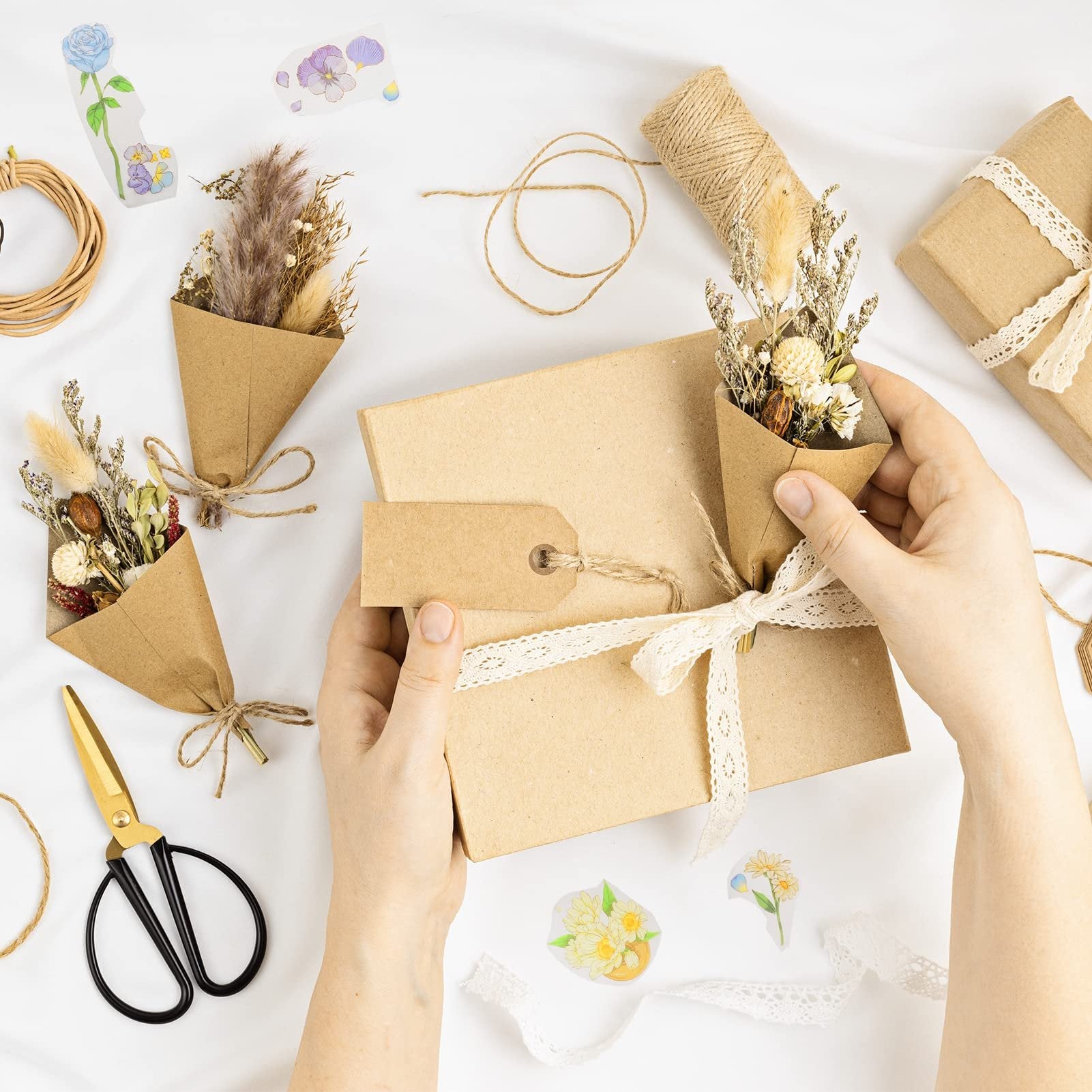 Hands wrapping a gift box with lace ribbon and a mini dried flower bouquet, surrounded by craft supplies.