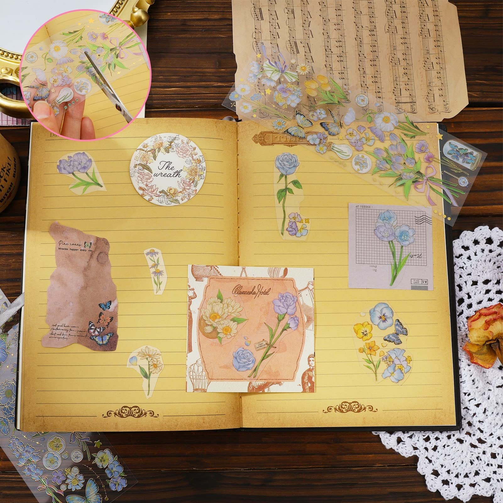Open journal with decorative stickers of flowers, butterflies, and a wreath applied to pages, with a hand cutting a sticker sheet in a circular inset.