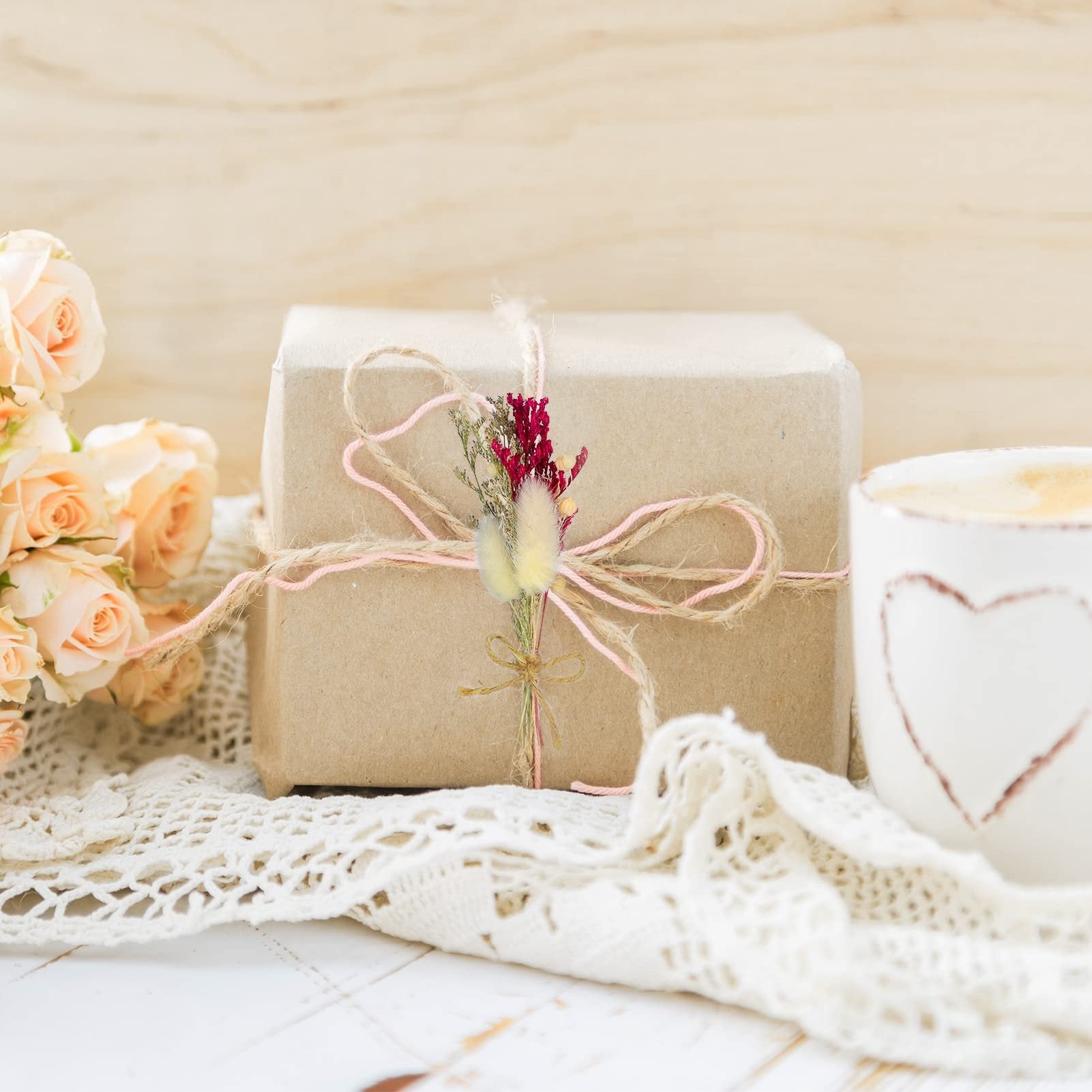 Gift box tied with twine and decorated with dried flowers, next to a bouquet of peach roses and a coffee cup with a heart design.
