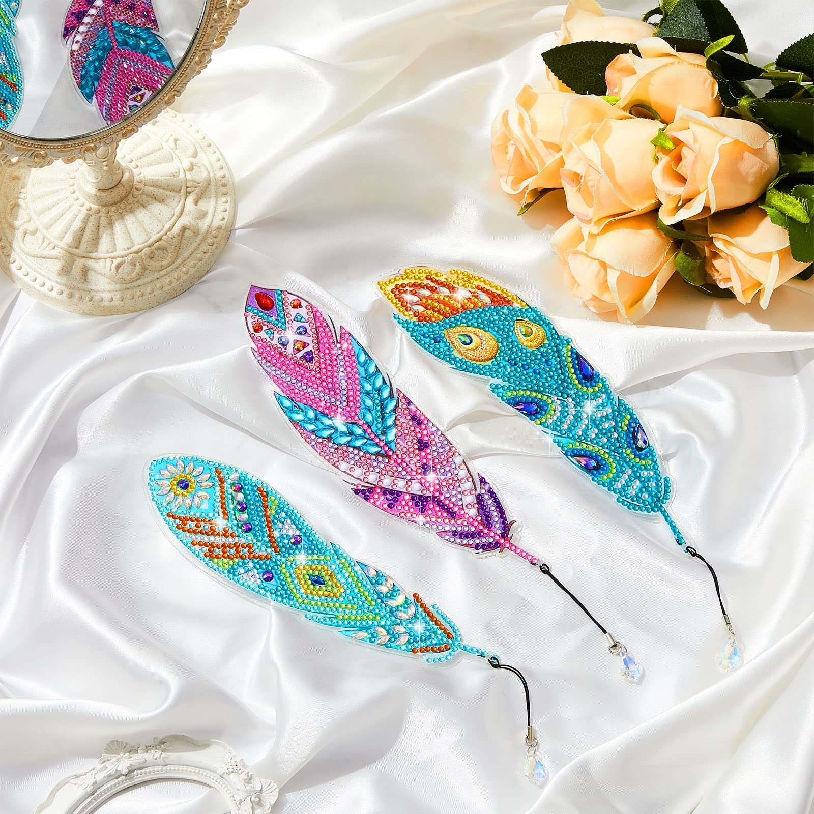 Three diamond-painted feather bookmarks with crystal pendants displayed on white satin, with a mirror and roses in the background.