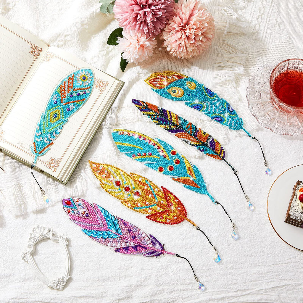 Diamond painting feather bookmarks laid out on a white cloth next to an open book and a glass of red liquid.