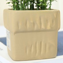 Close-up of a beige ceramic pot with artificial green plants, showing textured sides and a label.