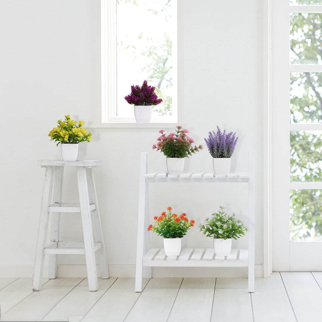 Six artificial potted plants in white pots arranged on a shelf and stool near a window.