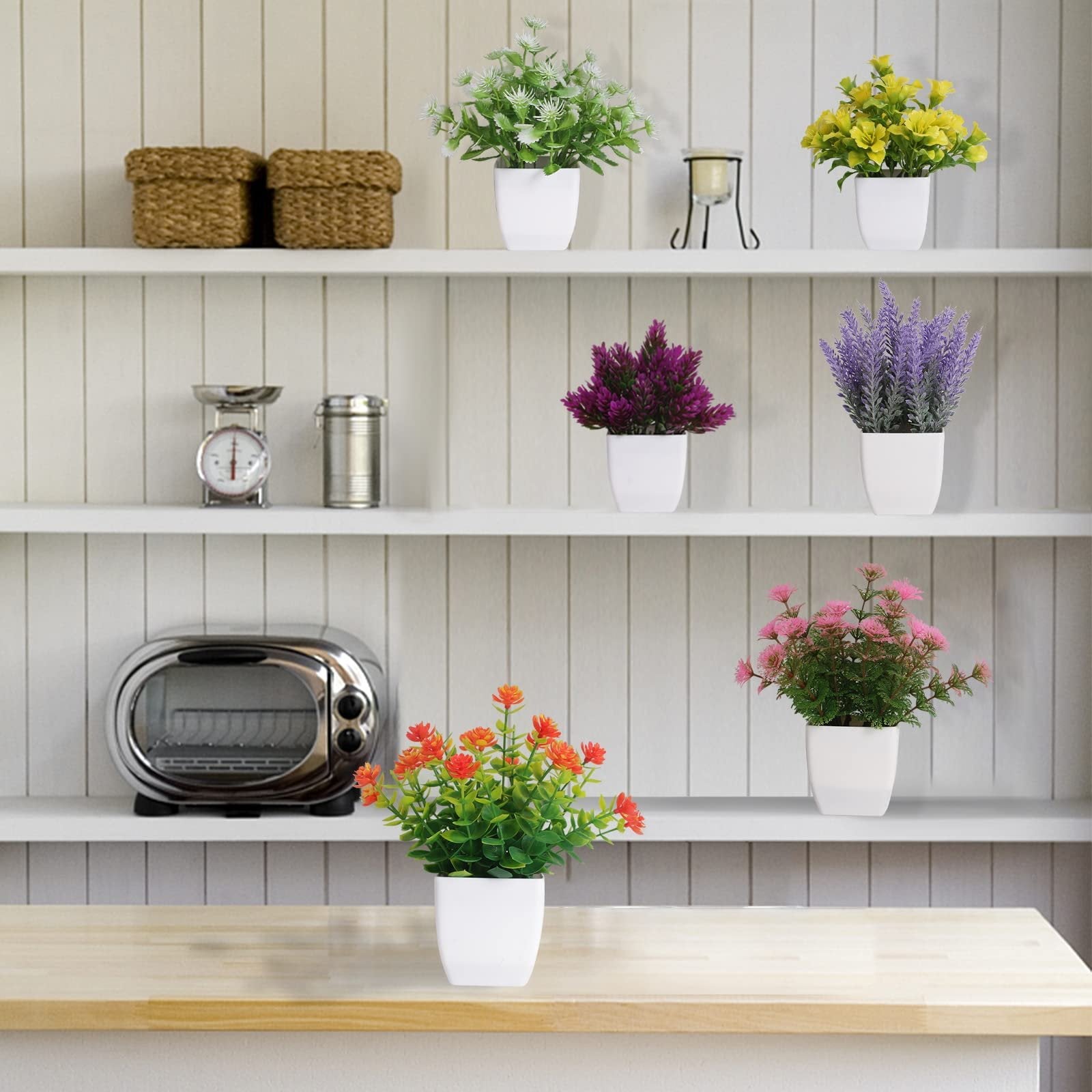 Six artificial potted plants in white pots arranged on shelves with decorative items and a toaster