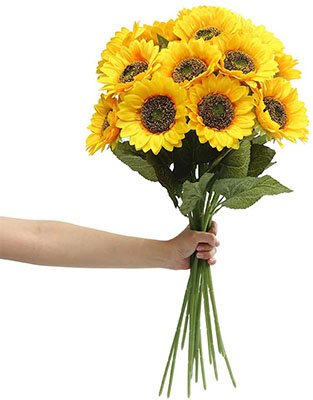 Hand holding a bouquet of six artificial sunflowers with green stems and leaves against a white background.