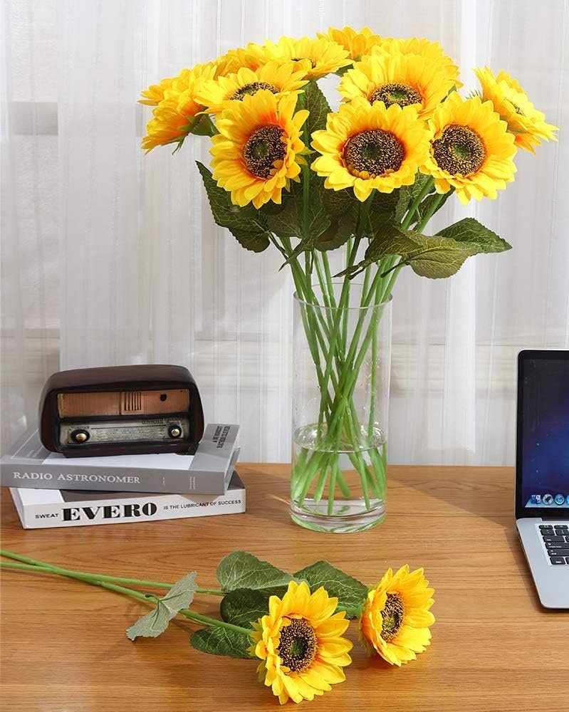 Six artificial sunflower stems in a glass vase on a wooden desk, with two stems in the foreground.
