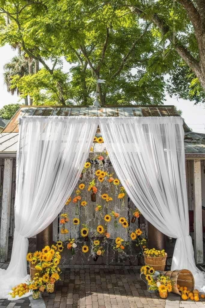 Wedding arch decorated with artificial sunflowers and white sheer curtains, with baskets of flowers and fruit at the base.