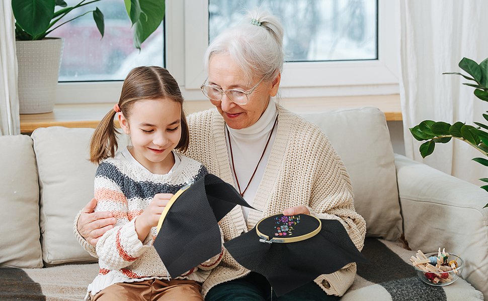 Grandmother and granddaughter doing cross-stitch on a couch, with a bowl of threads and needles nearby.