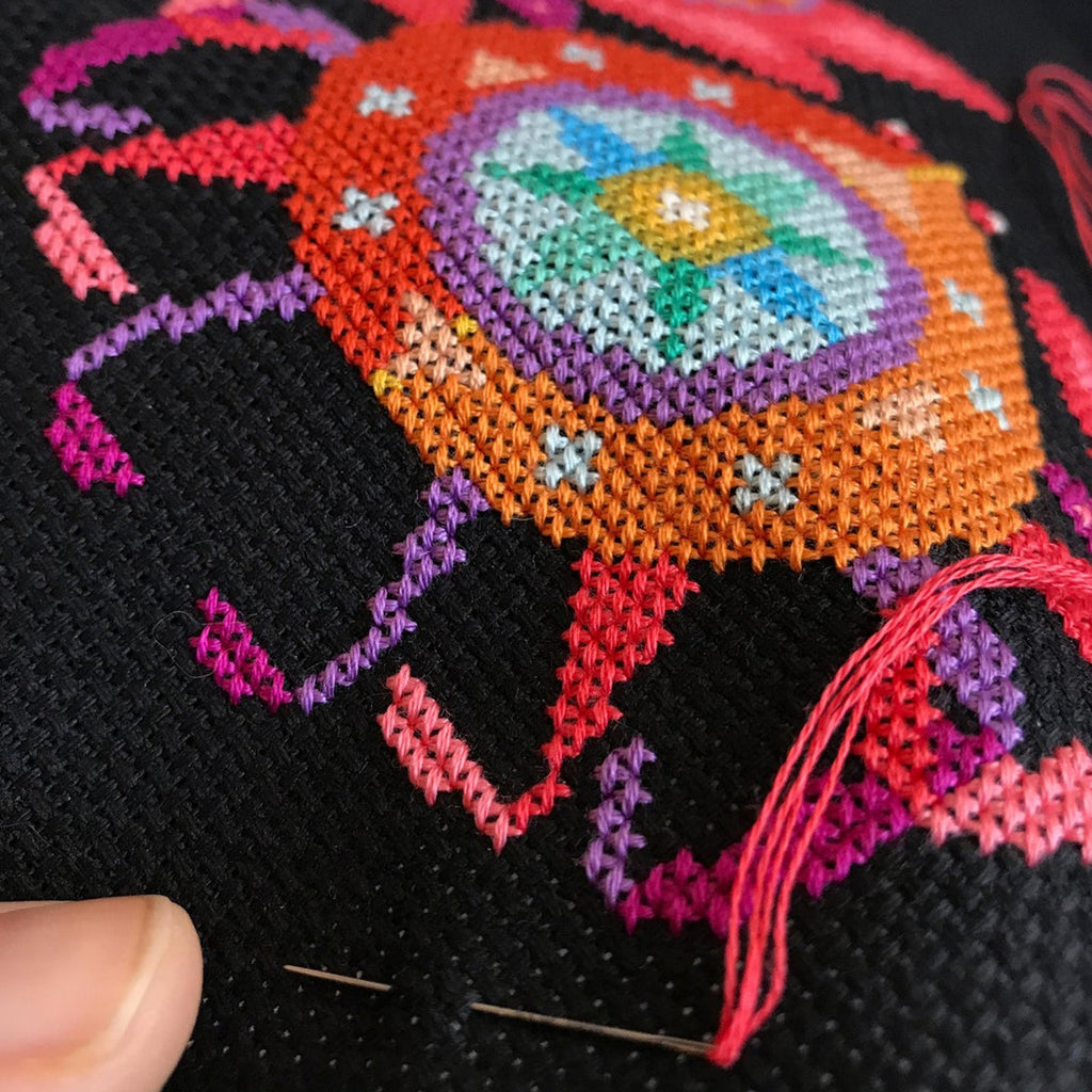 Close-up of black Aida cloth with colorful cross-stitch embroidery in progress, showing needle and thread.