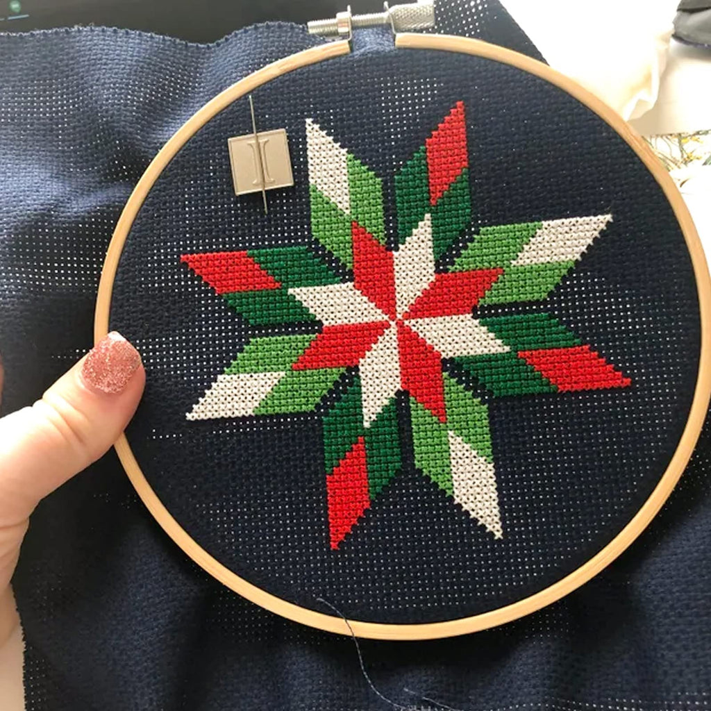 Cross stitch hoop with a partially completed red, green, and white star pattern on dark blue Aida cloth. A needle and a small square needle minder are visible.