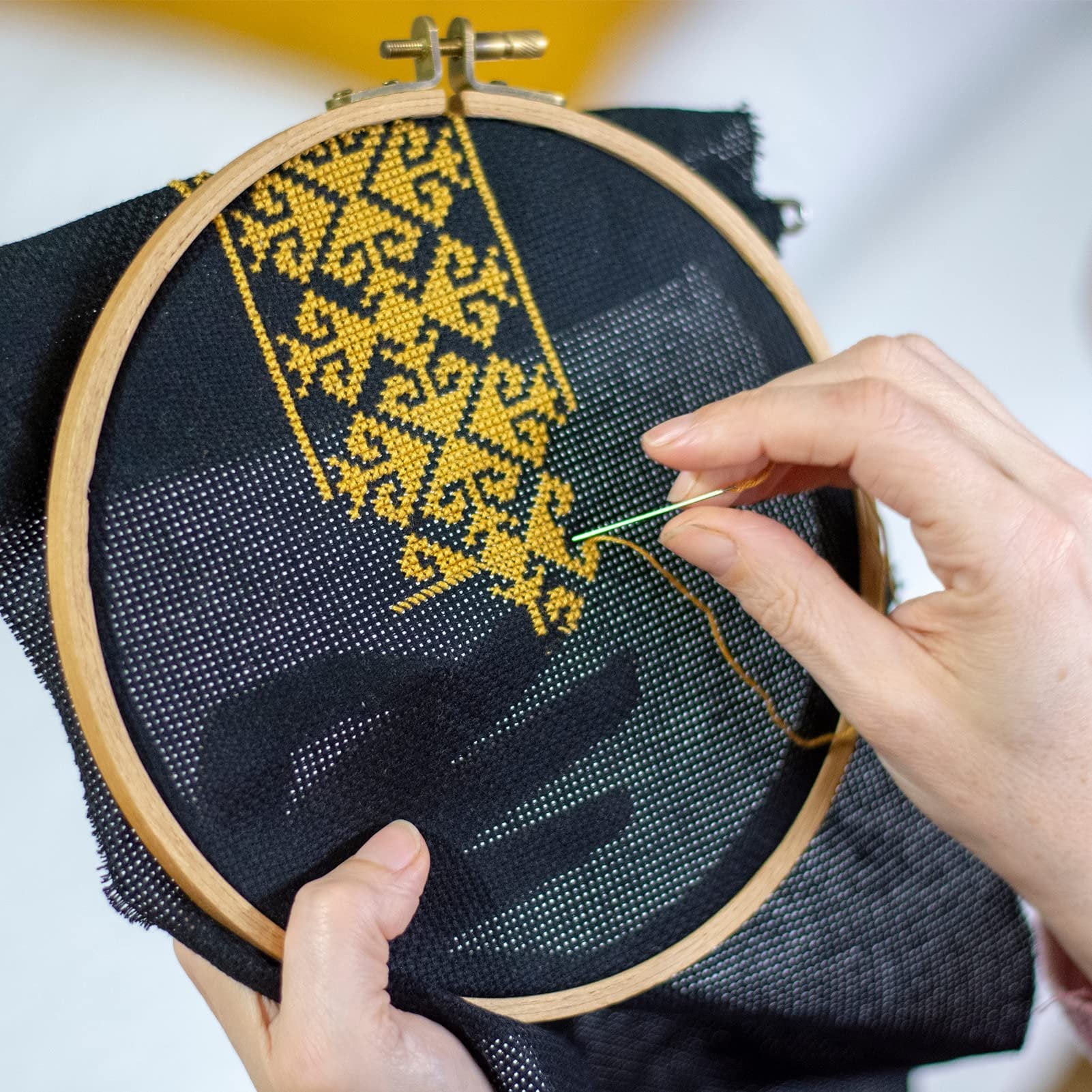 Hands cross-stitching a gold pattern onto black Aida cloth held in a wooden hoop