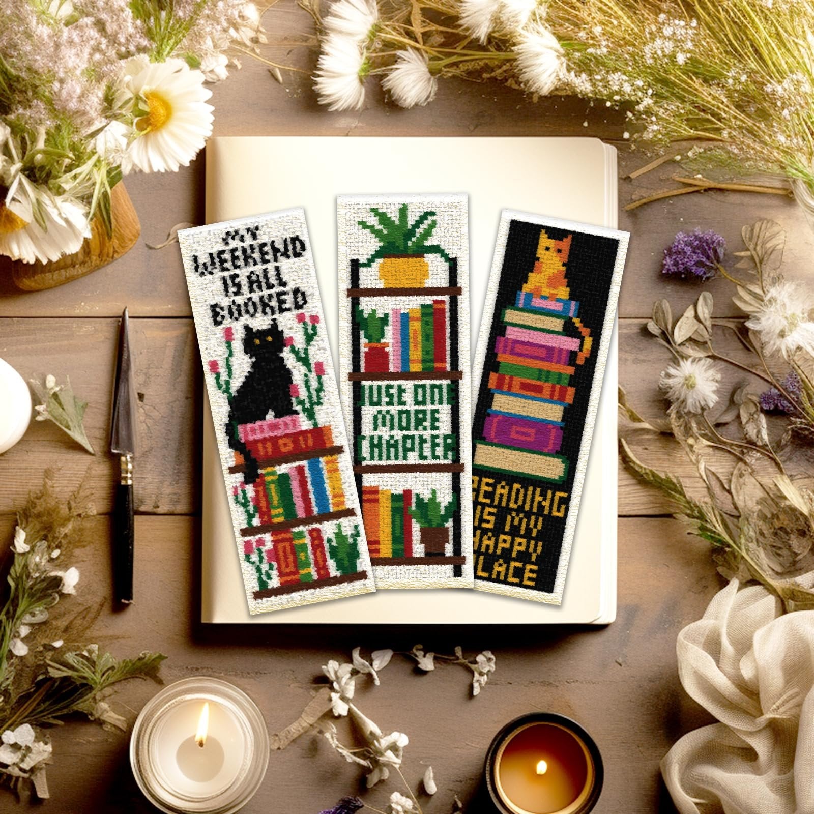 Three cross stitch bookmarks with book and cat designs, displayed on a book with decorative elements.