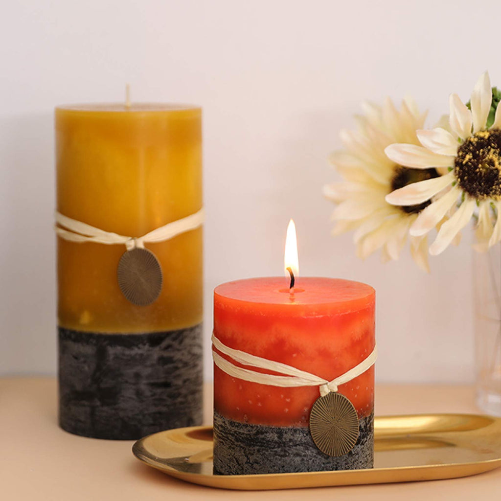 Two decorative pillar candles, one orange and one yellow, with raffia ribbon and metal charms, next to a vase of flowers.