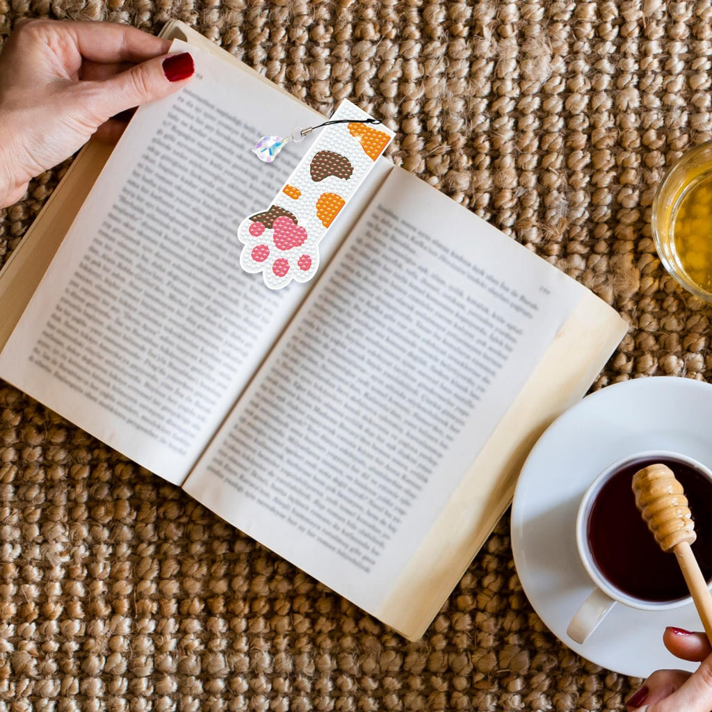Diamond painting cat paw bookmark with crystal pendant resting on an open book next to a cup of tea.