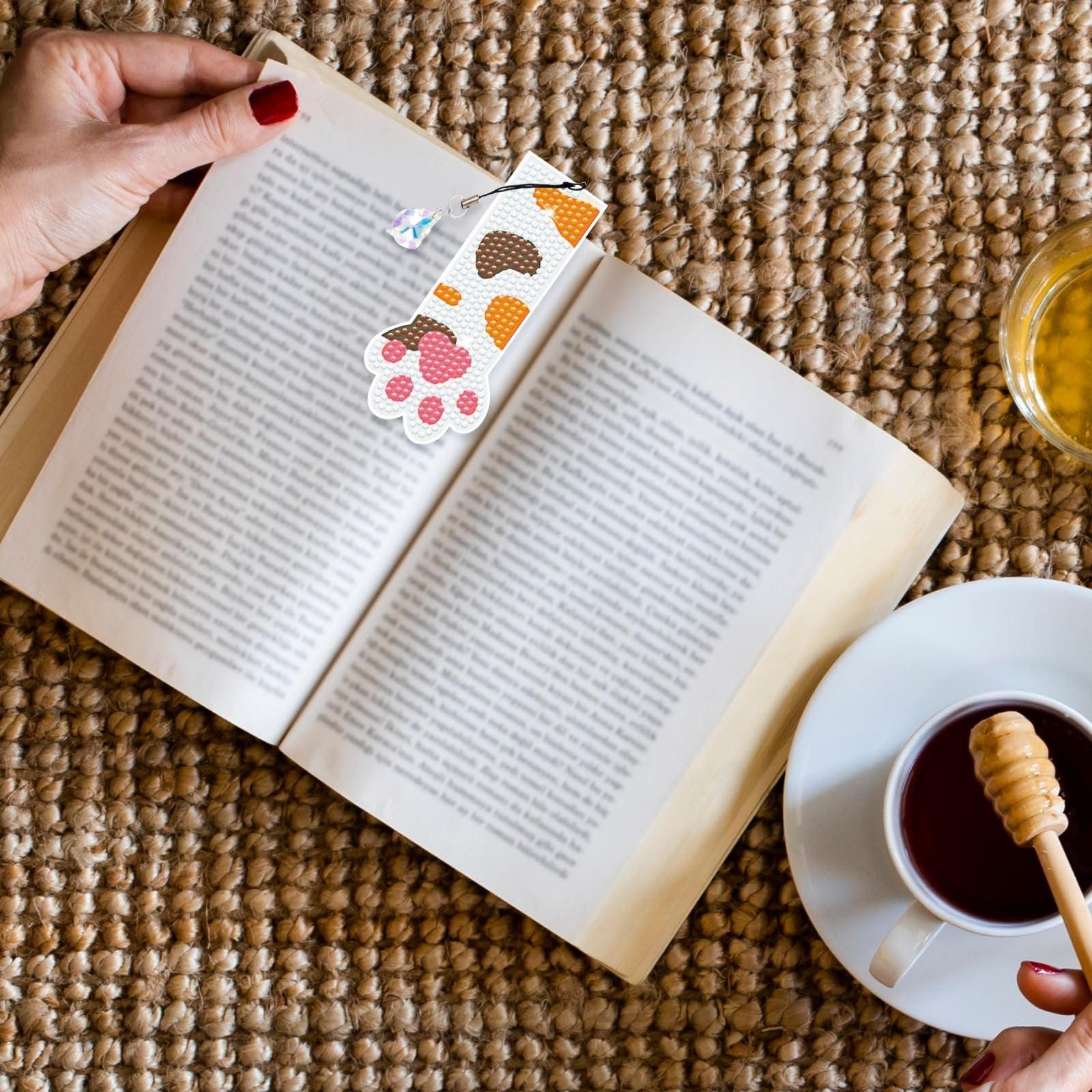 Diamond painting cat paw bookmark with crystal pendant resting on an open book next to a cup of tea.