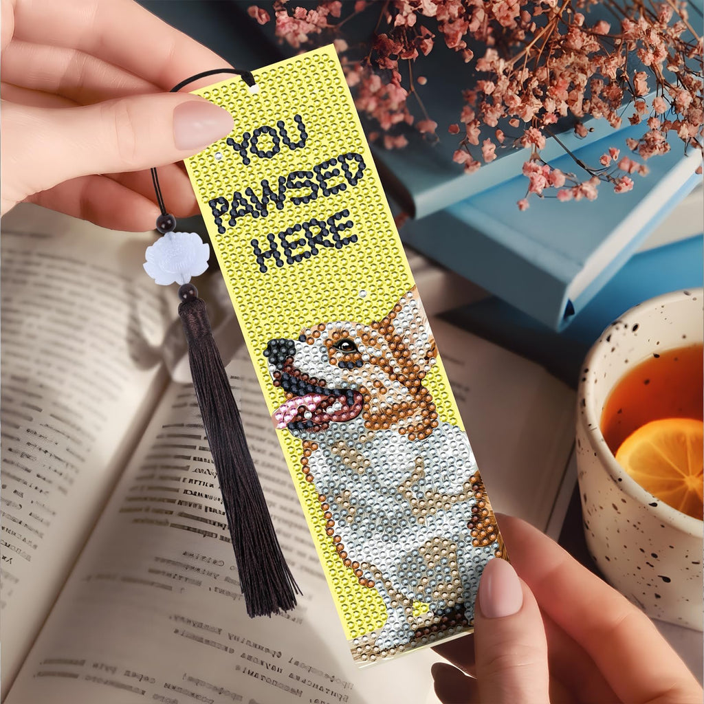 Diamond art bookmark with corgi dog design and tassel, held by hands near a book and tea.