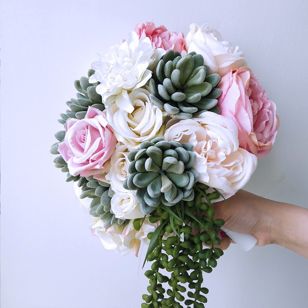 Hand holding a bouquet of artificial pink, white, and cream roses and succulents against a white background.