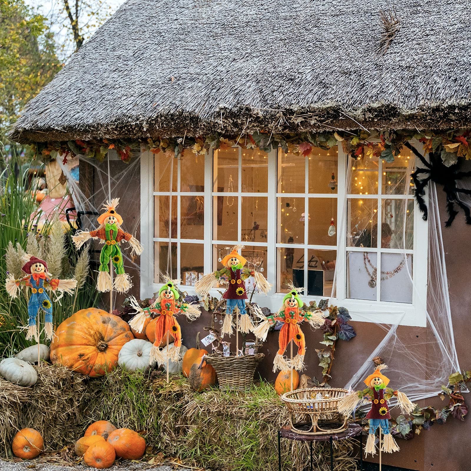 Six decorative scarecrows displayed with pumpkins and hay bales in front of a thatched-roof building for fall harvest and Halloween decor.