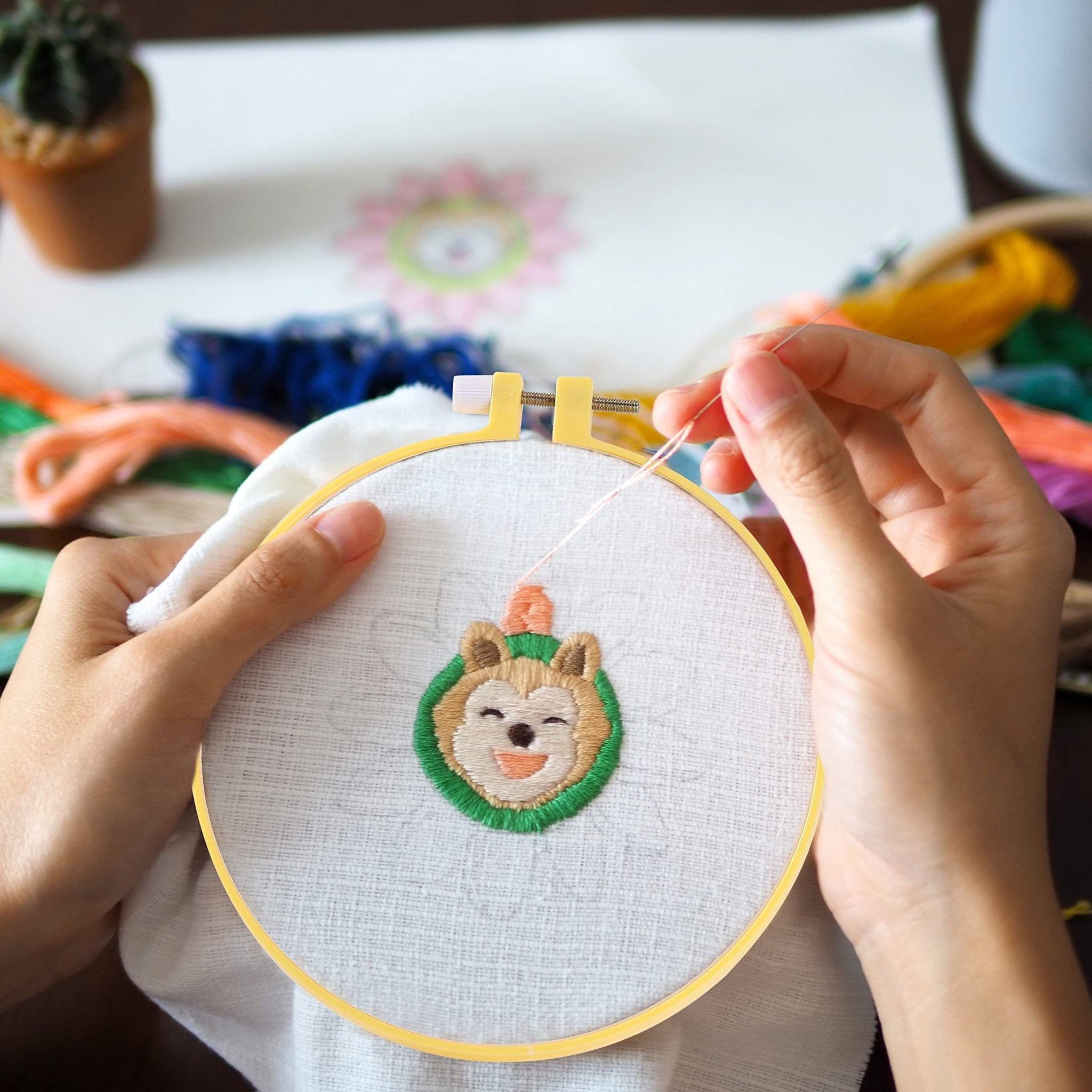 Hands holding an embroidery hoop with a partially stitched cartoon dog face design, surrounded by colorful threads and a small potted plant.