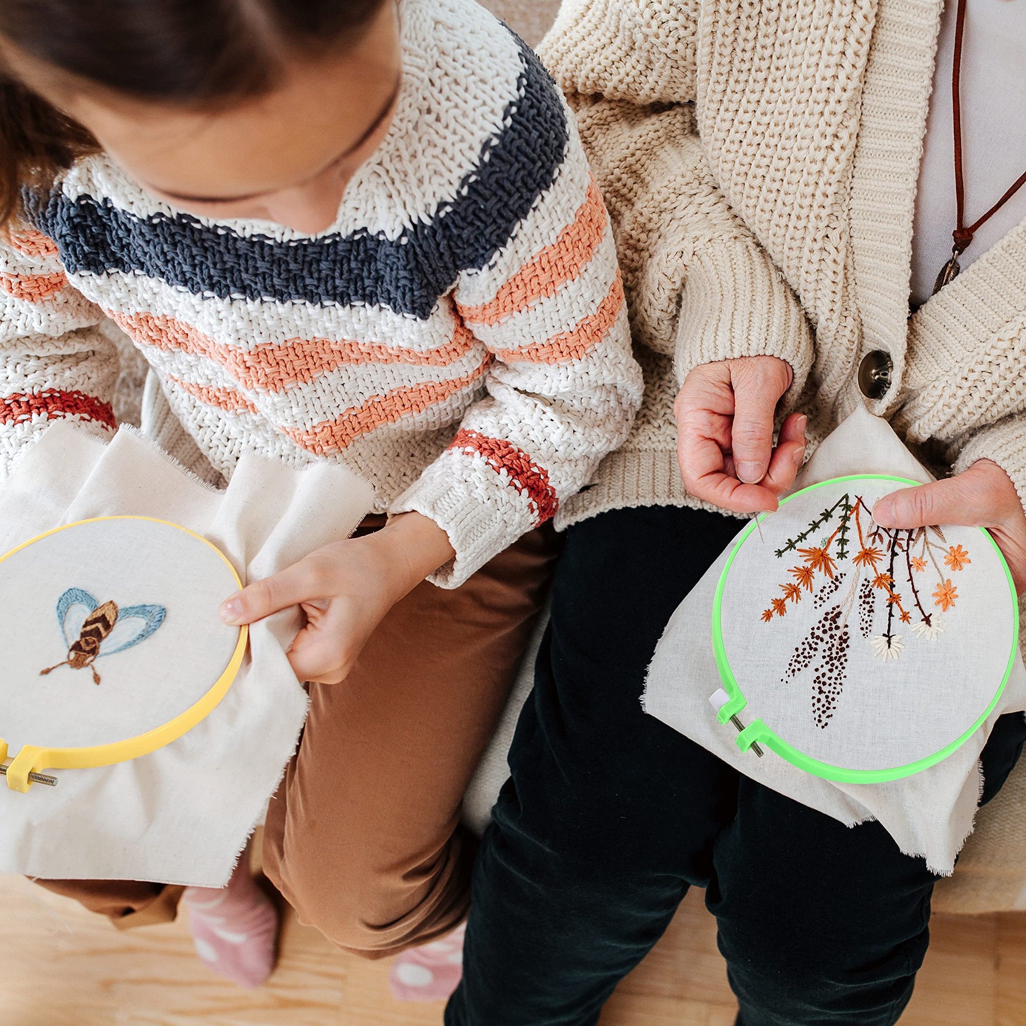 Close-up of a child and adult hands embroidering on fabric hoops, one with a bee design, the other with floral elements.