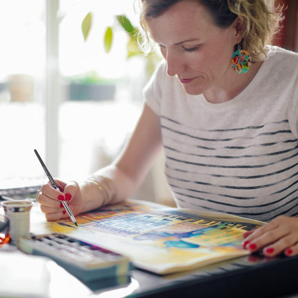 Woman painting with a round pointed tip brush and watercolor palette, close-up on hand and artwork