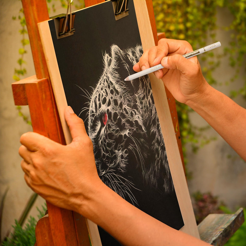 Artist's hands sketching a white charcoal leopard portrait on black paper with a white charcoal pencil.