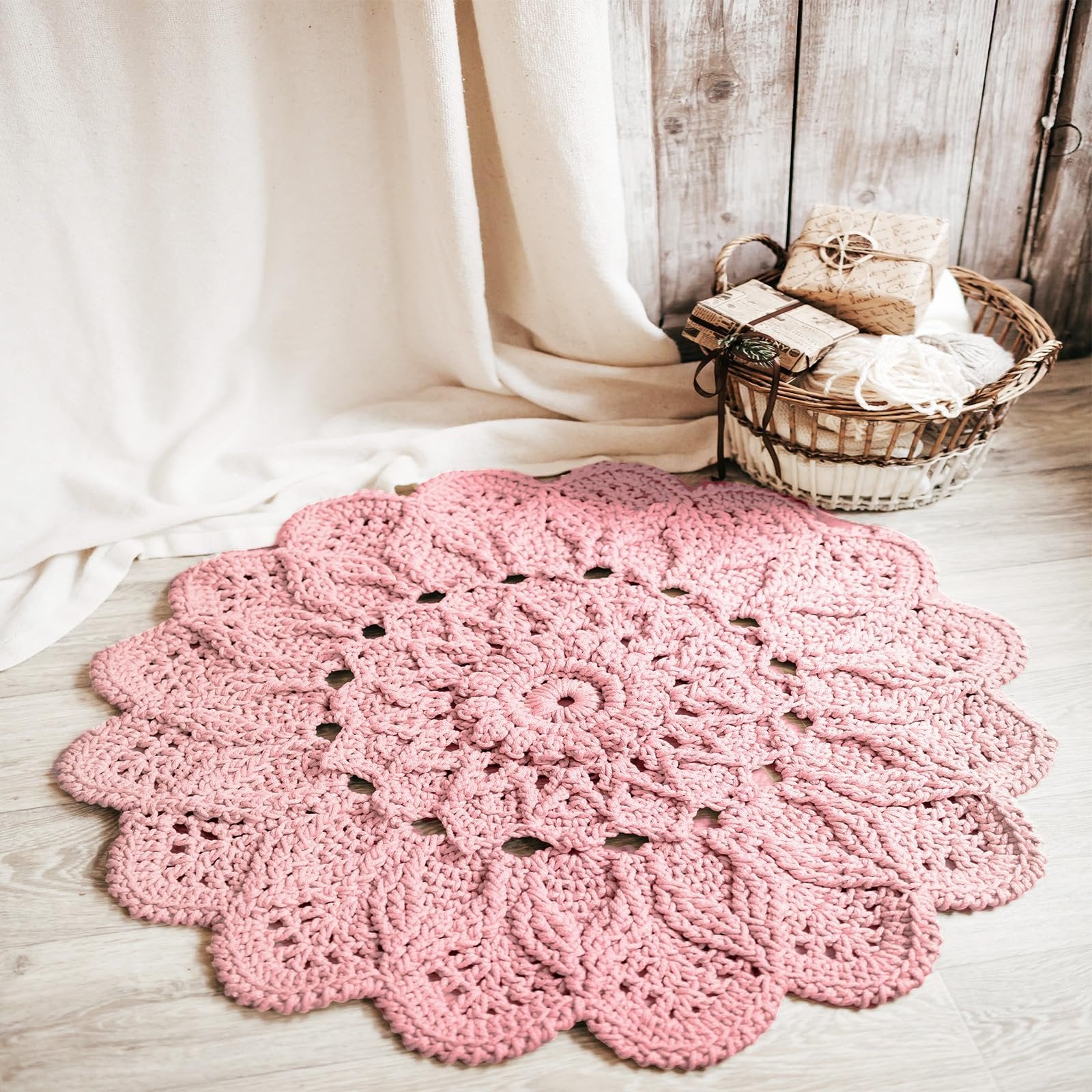 Pink crocheted doily with scalloped edges, displayed on a wooden floor with a basket of yarn and wrapped gifts in the background.