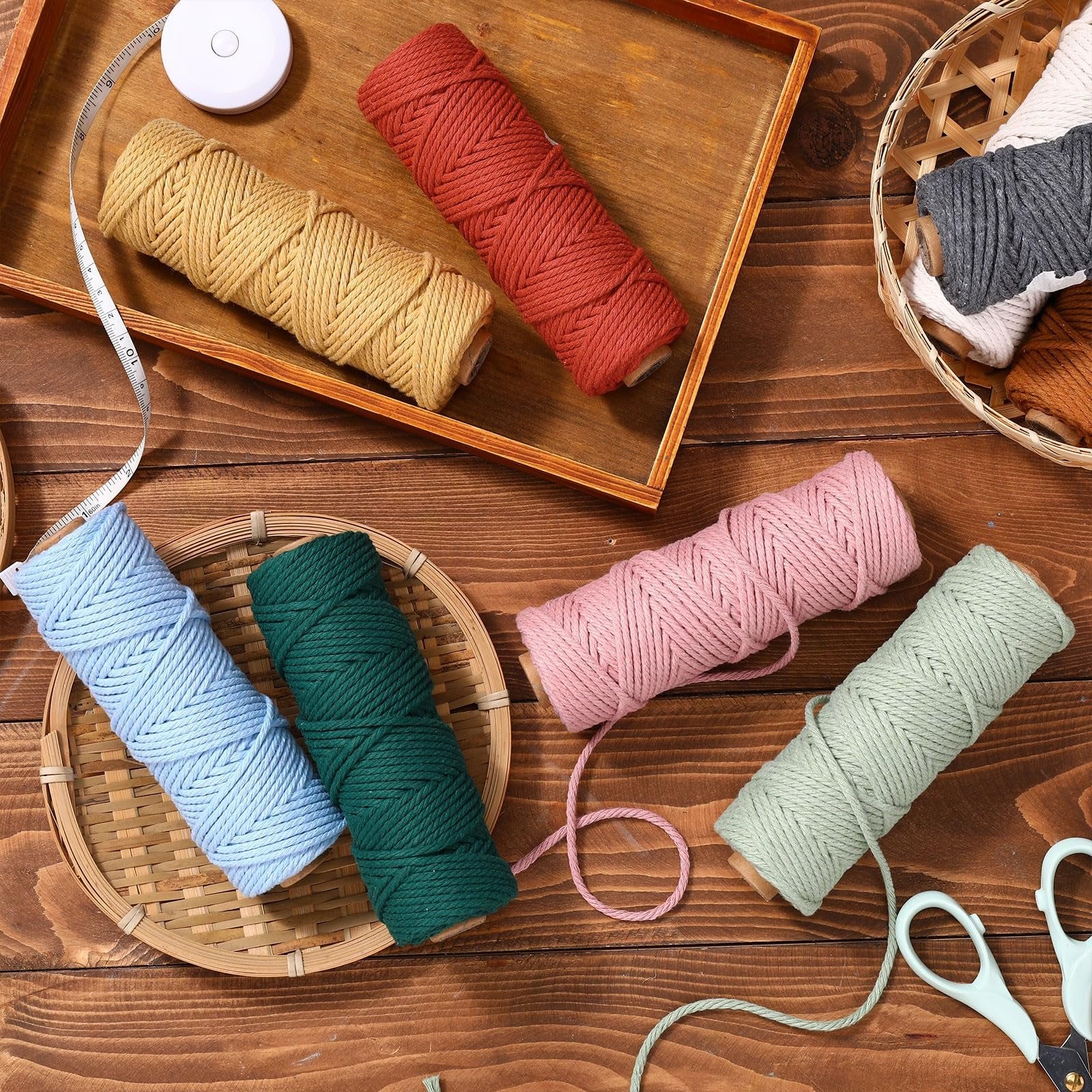 Six spools of 3mm cotton macrame cord in various colors arranged on a wooden surface with a tape measure and scissors.
