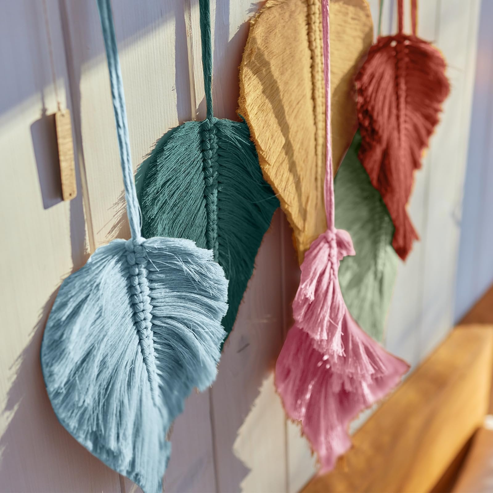 Close-up of macrame feather ornaments in blue, teal, mustard, and red hanging on a light-colored wooden surface.