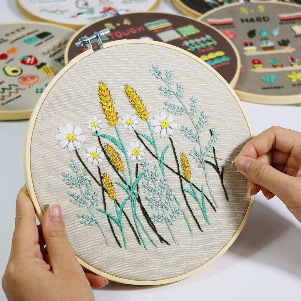Embroidery hoop with wheat and daisy pattern being stitched by hands, other patterned hoops in background
