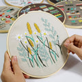 Embroidery hoop with wheat and daisy pattern being stitched by hands, other patterned hoops in background