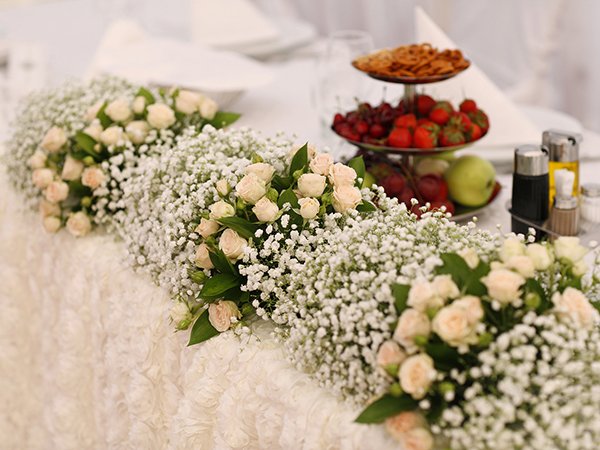 White artificial baby's breath and pale pink roses arranged along a table with fruit and condiments in the background.