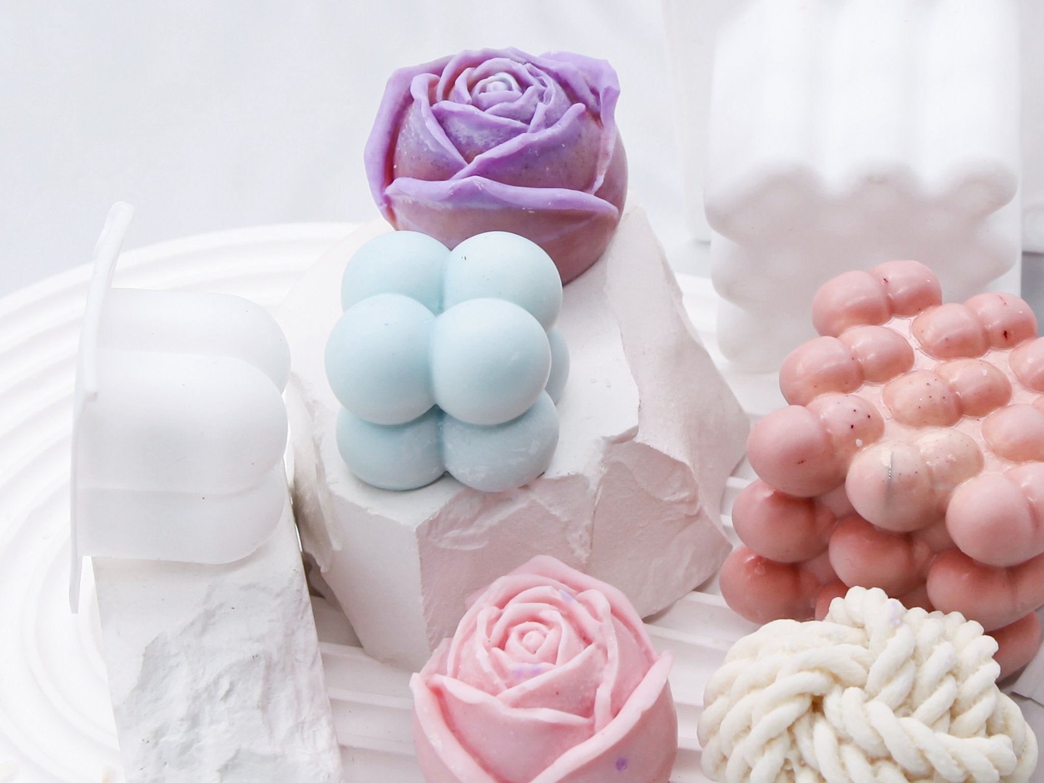 Assortment of colorful silicone candle molds in various shapes including roses, bubbles, and a braided design, arranged on white textured surfaces.