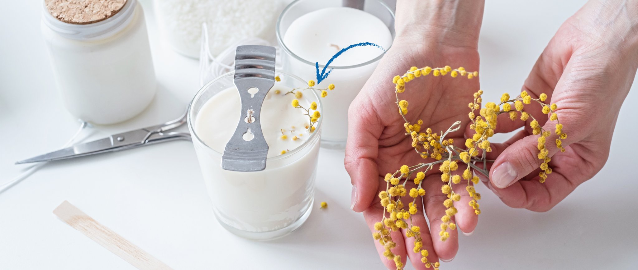 Hands decorating a candle with yellow flowers, surrounded by candle-making supplies like wax and wicks.