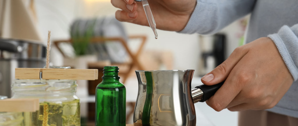 Close-up of hands preparing candle making with dropper adding liquid to pot and wick centered in jar